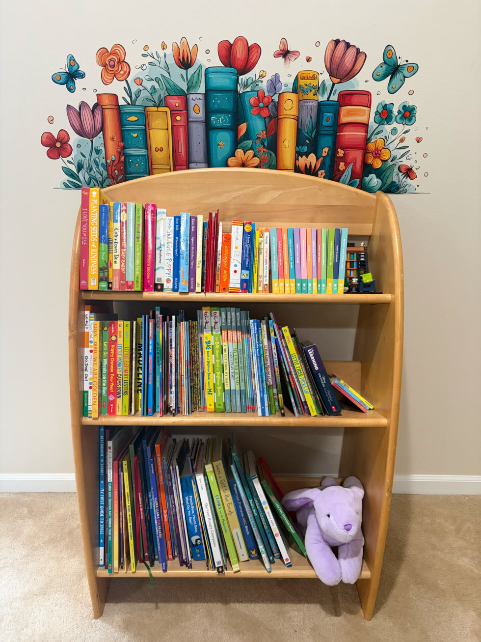 A children’s bookshelf under a printed band of illustrated books, flowers, and butterflies, from Bear Creek Murals.